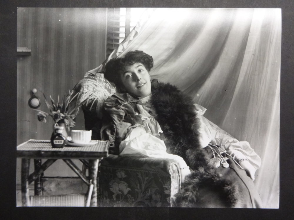 A black and white photograph of a woman sitting in a chair, wearing a boa with flowers and a teacup on a table next to her.