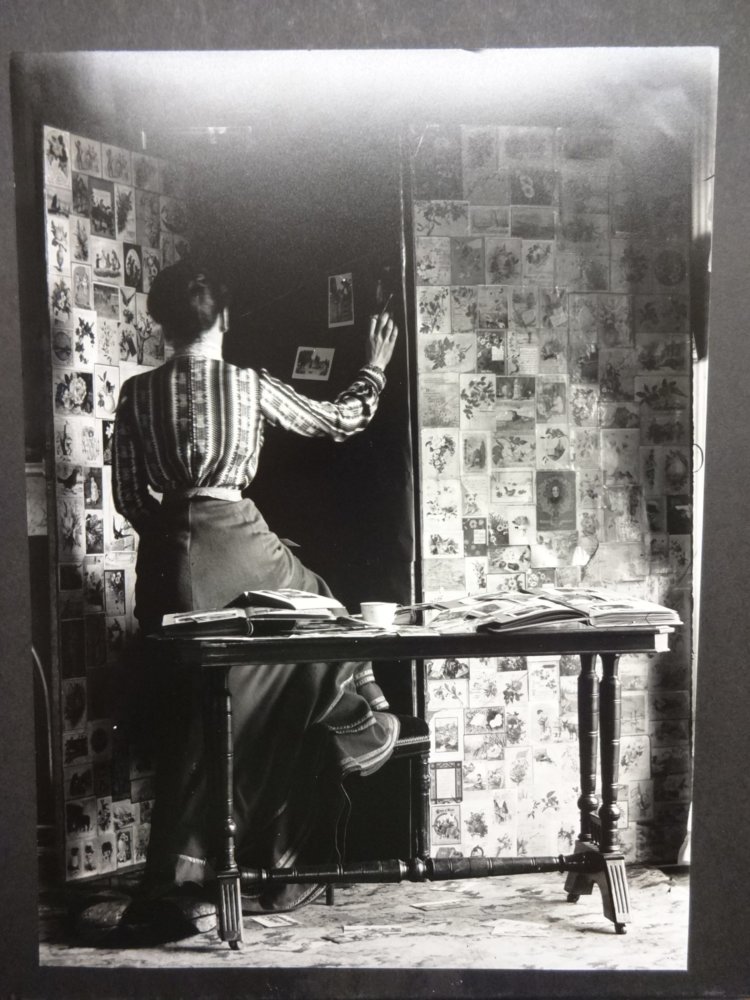 A black and white photograph of a woman standing with her back to the image, arranging photographs on a screen.