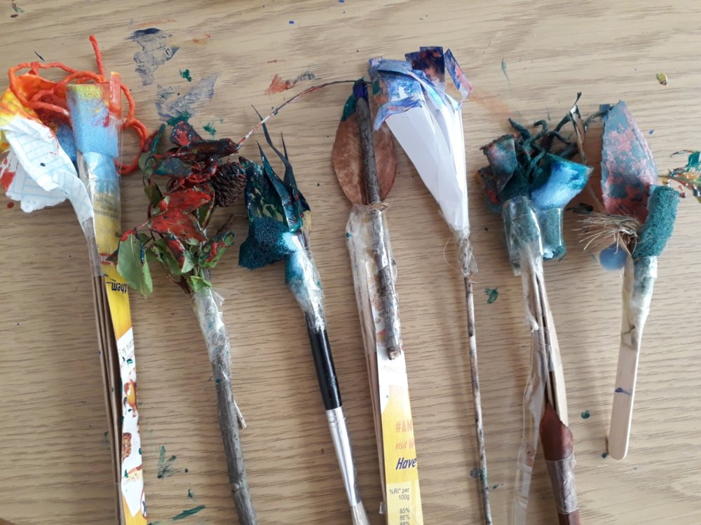 Seven used paintbrushes lined up on a table. They are made from different materials such as string and sponge.